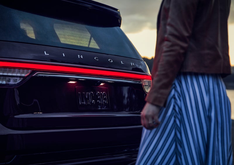 A person is shown near the rear of a 2024 Lincoln Aviator® SUV as the Lincoln Embrace illuminates the rear lights | Krenzen Lincoln in Duluth MN