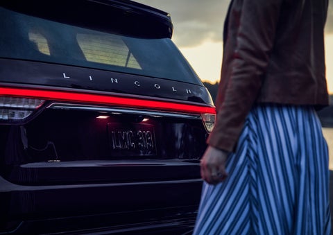 A person is shown near the rear of a 2024 Lincoln Aviator® SUV as the Lincoln Embrace illuminates the rear lights | Krenzen Lincoln in Duluth MN