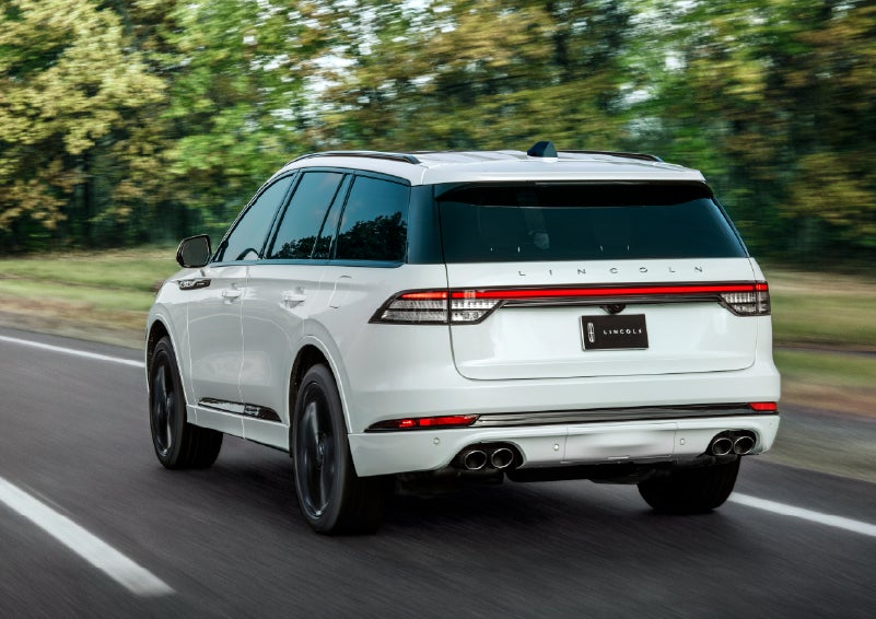 A white 2026 Lincoln Aviator® SUV is being driven away down a road lined with colorful trees. | Krenzen Lincoln in Duluth MN