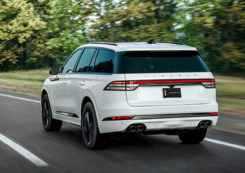 A white 2026 Lincoln Aviator® SUV is being driven away down a road lined with colorful trees. | Krenzen Lincoln in Duluth MN